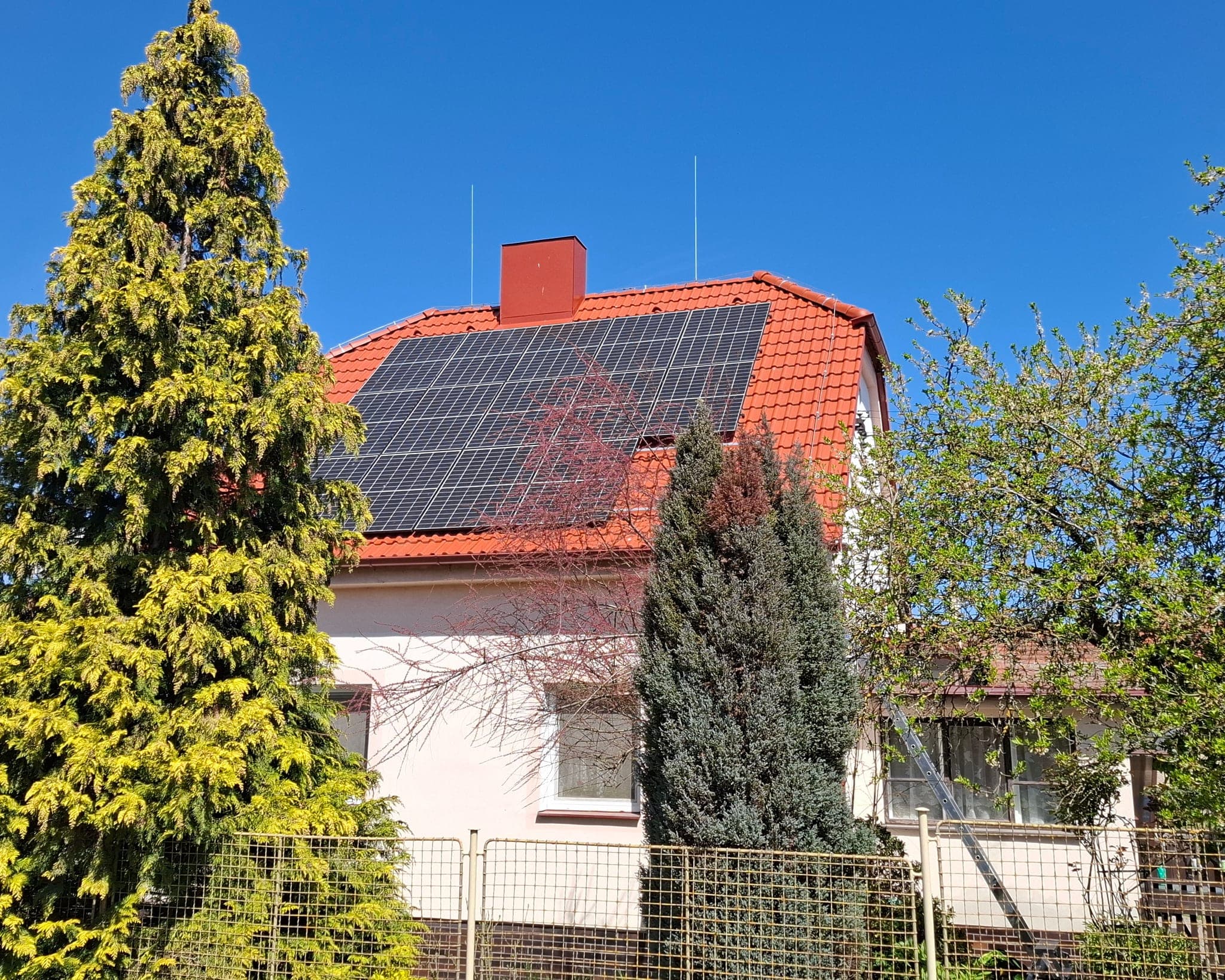 Rodinný dům s červenou střechou, na hřebeni dvě jímací tyče hromosvodu, pod nimi pole fotovoltaických panelů, Střelské Hoštice