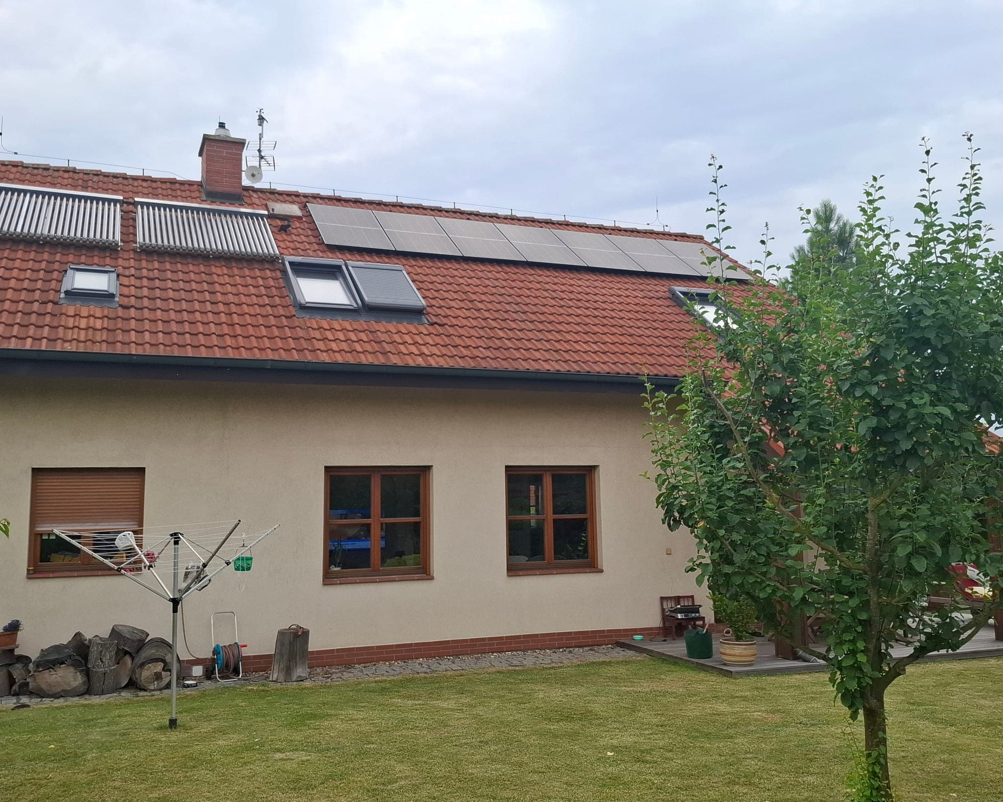 Fotovoltaické panely a střešní okna na červené střeše rodinného domu, před domem zahrada se stromem, Doksy